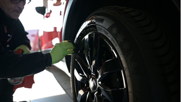 Person mit grünen Handschuhen reinigt einen schwarzen Reifen mit Felge in einer Werkstatt.