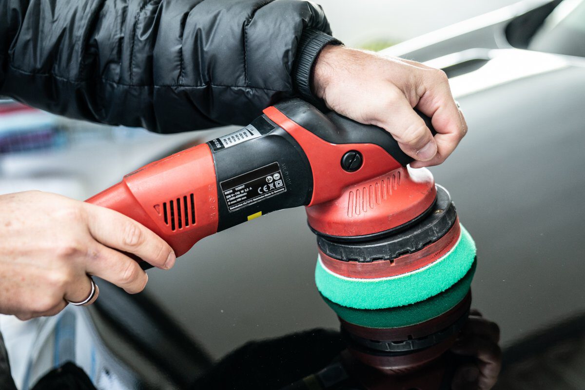 Eine Person benutzt eine Poliermaschine mit grünem Polierpad auf einer schwarzen Fahrzeugoberfläche.