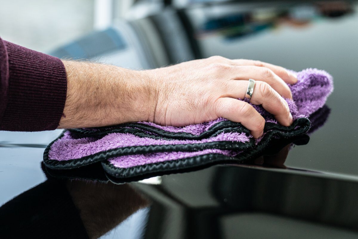 Eine Hand mit einem lila Mikrofasertuch, das über die Oberfläche eines schwarzen Autos wischt.