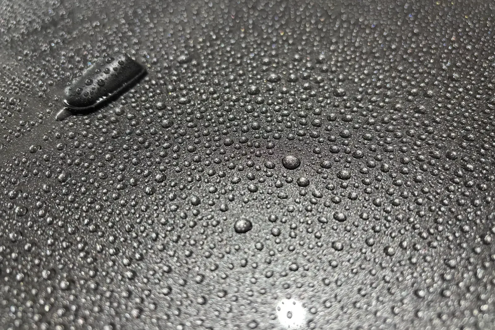 Wassertropfen auf einer glänzenden, dunklen Fahrzeugoberfläche mit reflektierenden Lichtpunkten.