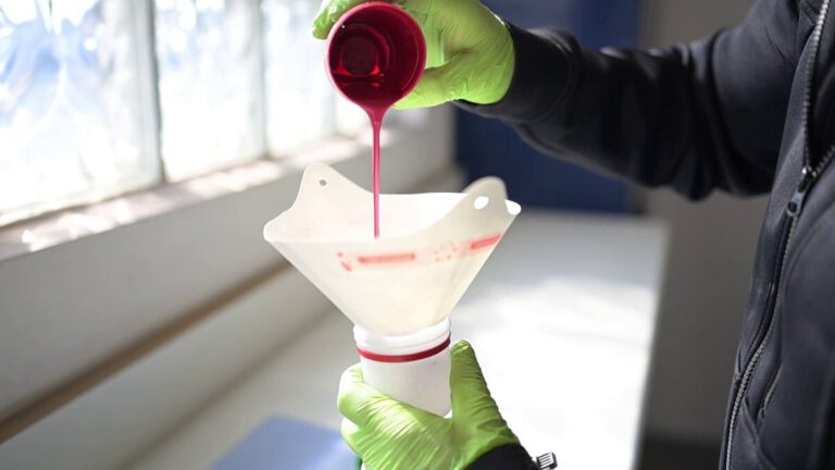 Person mit grünen Handschuhen, die roten Lack in einen Trichter gießt.