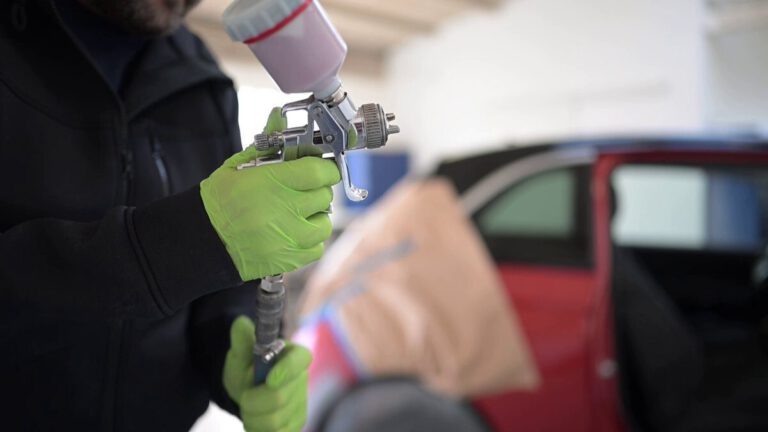 Ein Mann hält eine Lackierpistole mit grünen Handschuhen in einer Werkstatt für Fahrzeugaufbereitung.