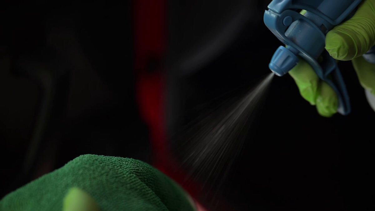 Eine Hand mit grünen Handschuhen sprüht Reinigungslösung aus einer blauen Sprühflasche auf ein grünes Tuch.