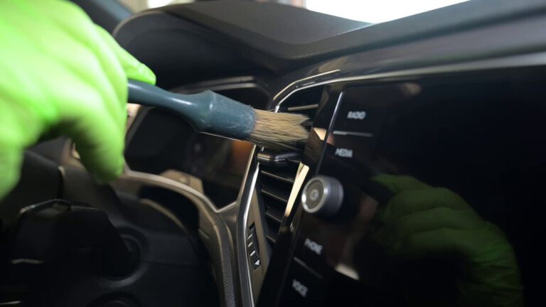 Eine Person mit grünen Handschuhen reinigt das Armaturenbrett eines Autos mit einem Pinsel.