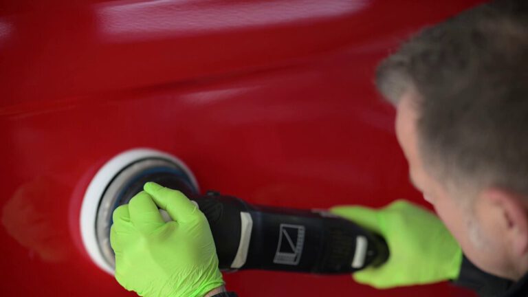 Ein Mann mit grünen Handschuhen benutzt eine Poliermaschine auf einer roten Fahrzeugoberfläche.