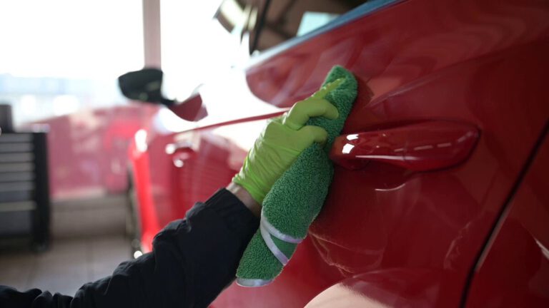 Eine Hand mit einem grünen Mikrofasertuch reinigt die Tür eines roten Autos.